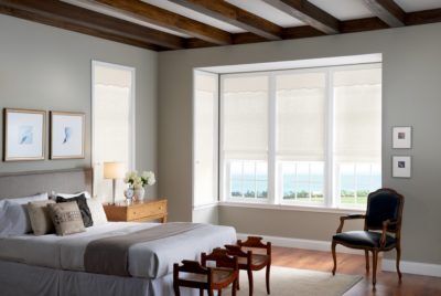 Partially sheer white shades half open on three vertical windows in a bedroom.